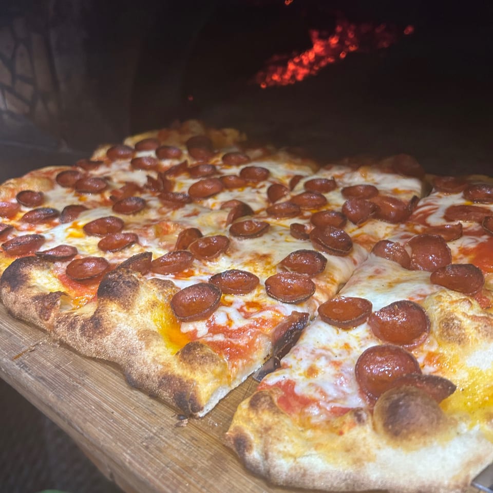 Pepperoni pizza with a crispy crust on a wooden peel, with a fiery oven in the background.