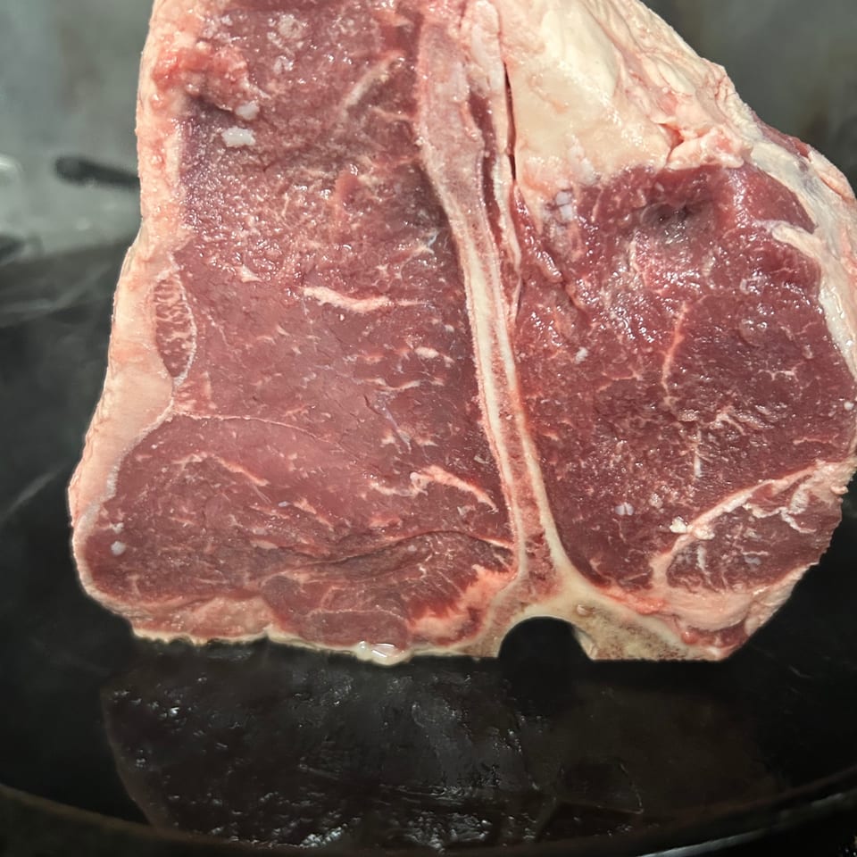 A raw, thick-cut T-bone steak with visible marbling sits in a dark pan.