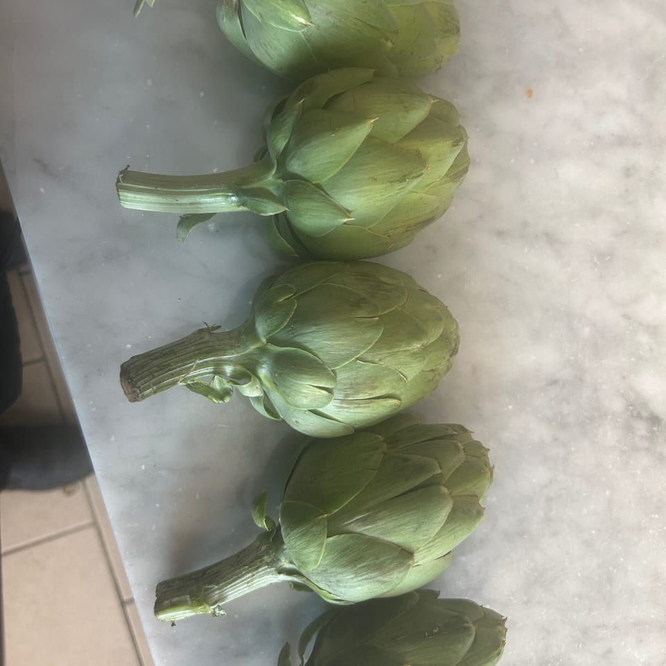 Five fresh green artichokes lined up on a white marble surface, ready for cooking.