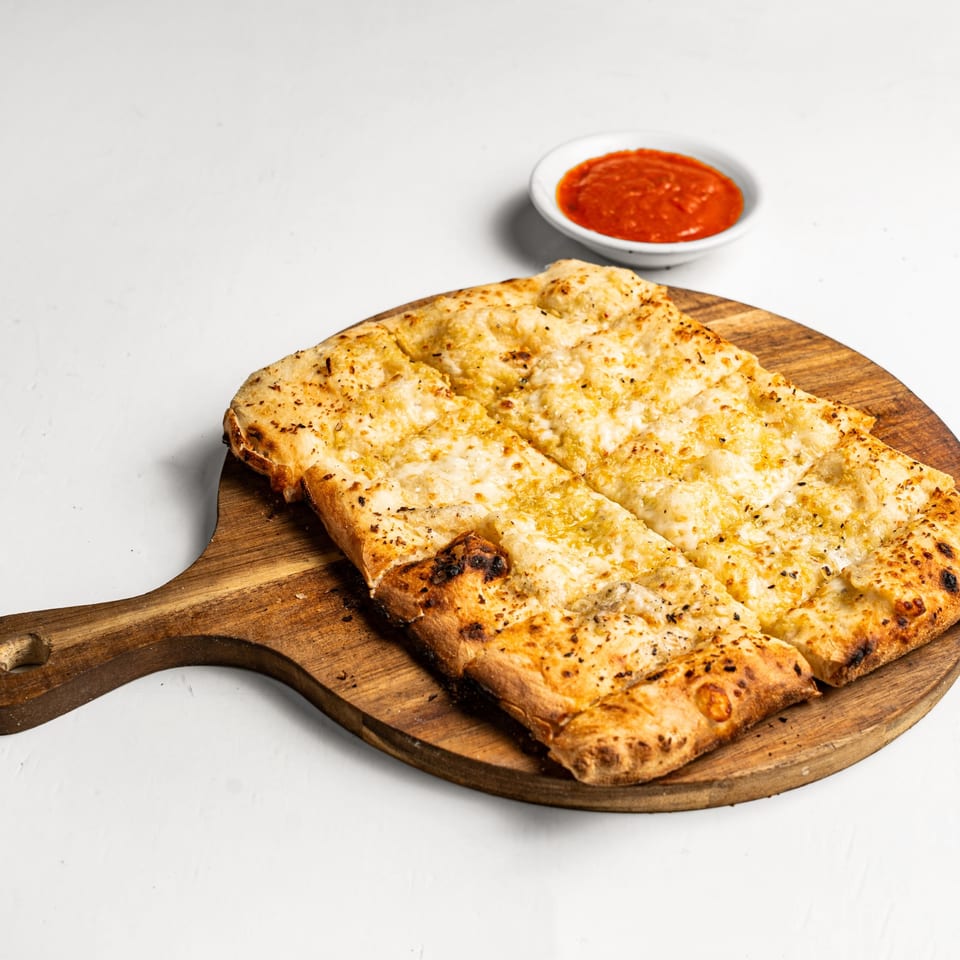 Cheesy garlic bread, cut into strips, on a wooden board with a side of red dipping sauce.