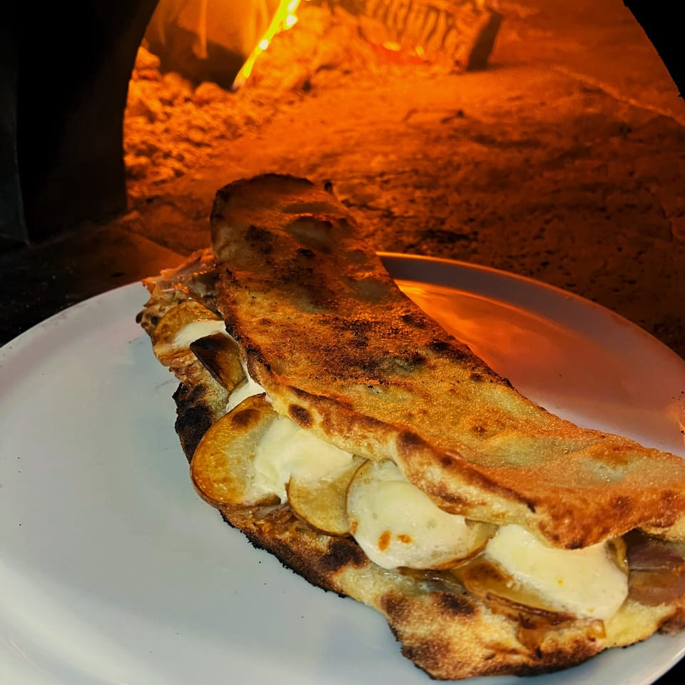 A calzone with melted cheese and potatoes on a white plate, with a wood-fired oven in the background.